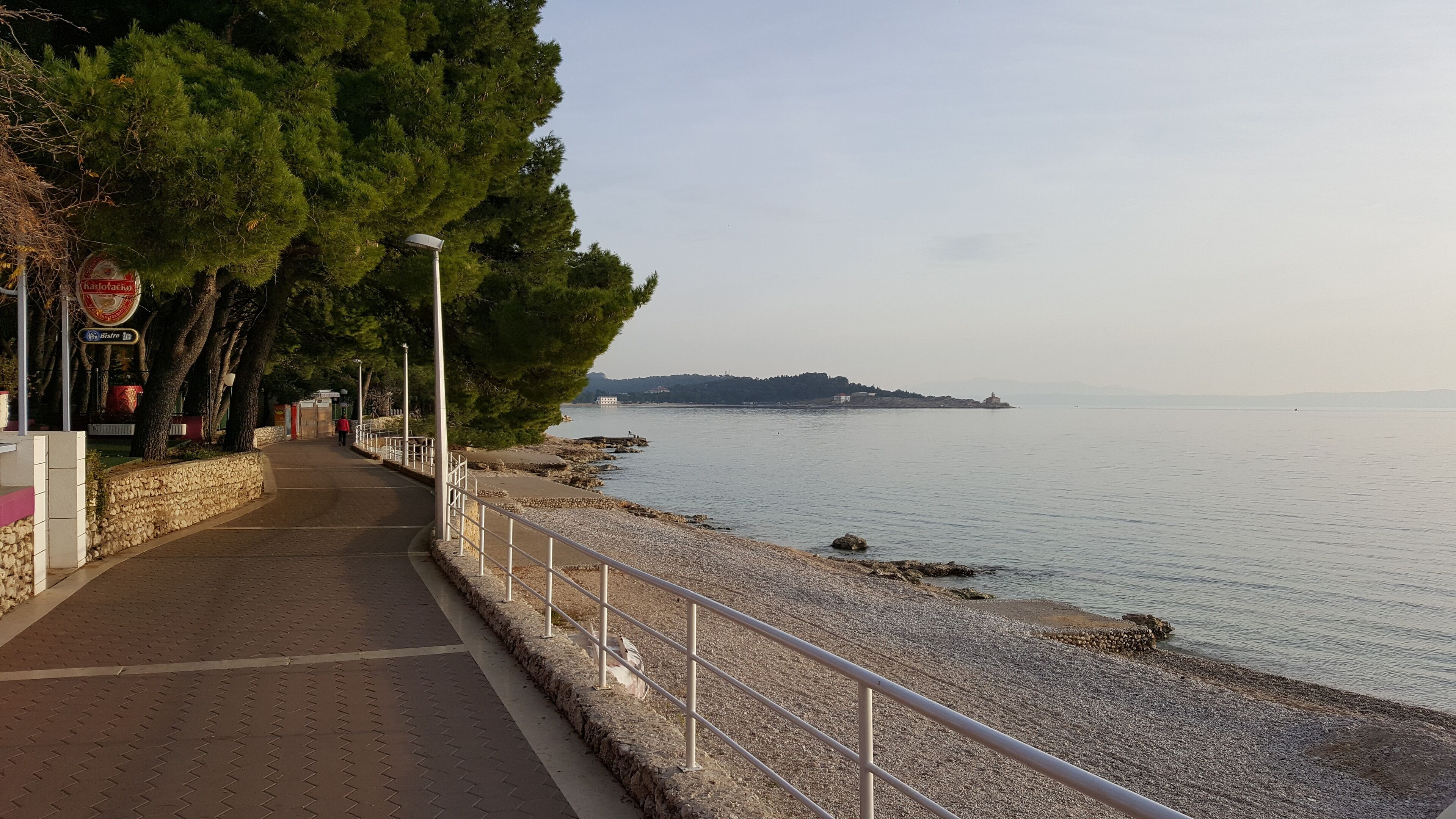Un chemin pavé longe un plan d’eau, avec une rambarde d’un côté et des arbres de l’autre.
