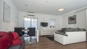 A modern living room with a white sofa, a red sofa, and a grey sofa. There is a dining area with a table and chairs. A TV is mounted on the wall, and there is a window with blinds.