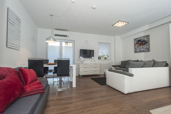 Un salon moderne avec un canapé blanc, un canapé rouge et un canapé gris. On y trouve un coin repas avec une table et des chaises. Un téléviseur est fixé au mur, et il y a une fenêtre avec des stores.