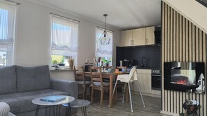 A modern living room with a grey sofa, a dining table with chairs, a small round table, and a kitchen area with wooden cabinets, a stove, and a grill. - 4 * FH with garden, playground, sauna, fitness, 2 bedrooms, kitchen with dishwasher (Ludorf)