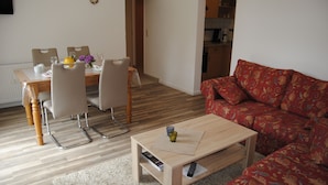 A living room with a wooden coffee table, a red patterned sofa, a dining table with chairs, and a kitchen area in the background. - Vacation apartment directly by the dike (Niedersachsen)