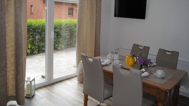 A dining area with a table set for four, a television on the wall, and a view of a brick building and greenery outside. - Vacation apartment directly by the dike (Niedersachsen)