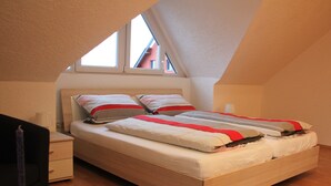 A bedroom with a sloped ceiling, a bed with striped bedding, a bedside table, and a window with a view of a building. - 3-star vacation apartment on the Rheinsteig with a magnificent view over the Rhine Valley (Rüdesheim am Rhein)