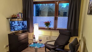A living room with a TV, a table with wine glasses, a chair, and a window with curtains. - Beautiful apartment in a central location in Wedel (Hamburg) (Wedel)