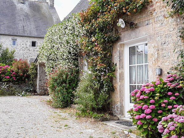 A stone building with a white door and window, surrounded by a gravel driveway and lush greenery.