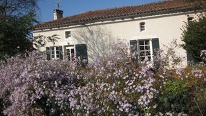 Un bâtiment blanc, doté d’un toit en tuiles et de volets verts, entouré de plantes en fleurs.