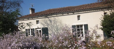 A white building with a tiled roof and green shutters, surrounded by flowering plants.
