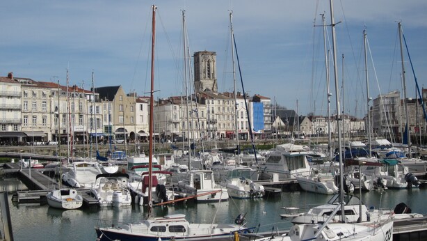 Un port de plaisance où sont amarrés différents types de bateaux, un bâtiment historique en arrière-plan et un ciel dégagé.