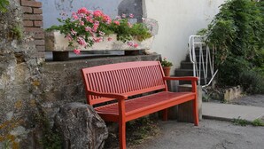 Un banc rouge avec un jardinier rempli de fleurs roses, devant un bâtiment aux volets rouges.