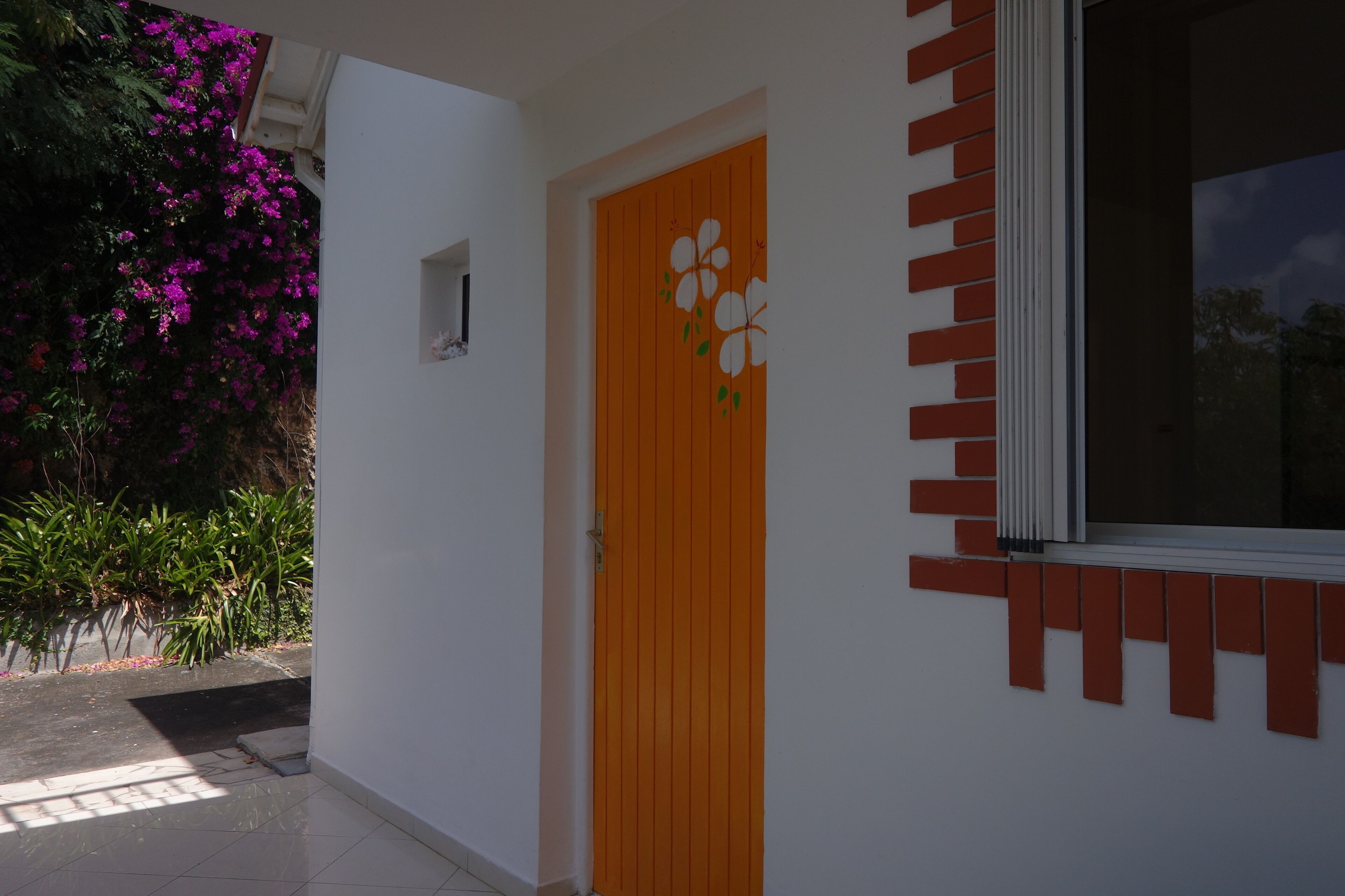 A white building with a wooden door featuring a floral design, a window with a red frame, and a brick accent wall.