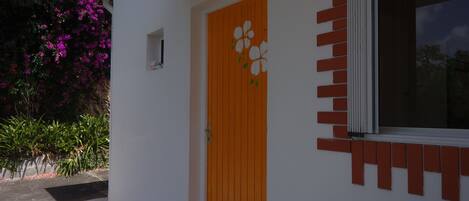 A white building with a wooden door featuring a floral design, a window with a red frame, and a brick accent wall.