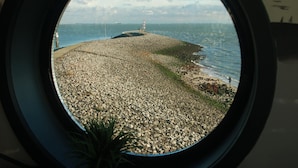 A view through a circular window of a rocky shoreline with a lighthouse in the distance. - Characteristic apartment, renovated, 500m from the sea, beach and town center (Vlissingen)