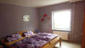 A bedroom with a wooden bed, purple floral bedding, a nightstand, a window with curtains, and a wall-mounted mirror.