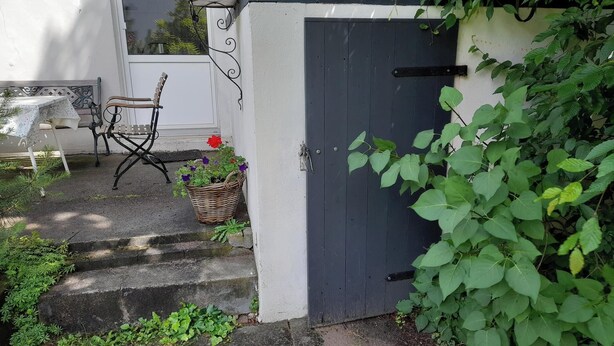 Entrada de jardí amb una porta de fusta fosca, una banca i una taula amb una cistella de flors.
