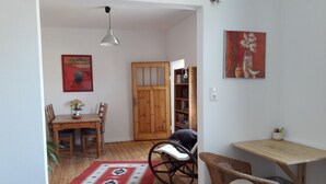 Una habitació amb taula de dinar de fusta, cadires, una catifa vermella amb dibuixos, una cadira de vimet i una estanteria, a més d'un quadre penjat a la paret.