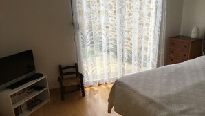 Una camera da letto con un letto, una TV su un mobile, una piccola sedia, un comò in legno e una finestra con tende leggere.
