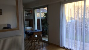 Una stanza con pavimento in legno, un tavolo con tovaglia, una sedia, un termosifone, una finestra con tende leggere e una porta che dà su un balcone.