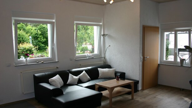 A modern living room with a black leather sofa, a wooden coffee table, and a radiator. There are two windows with views of greenery outside. - Detached holiday home, Apartment 2 / for 2-4 persons, W-LAn free (Mehren)