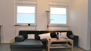 A modern living room with a black sofa, a wooden coffee table, and two windows with blinds. - Detached holiday home, Apartment 2 / for 2-4 persons, W-LAn free (Mehren)