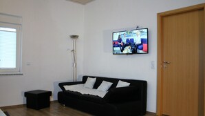 A living room with a black sofa, a television mounted on the wall, a wooden door, and a window with blinds. - Detached holiday home, Apartment 2 / for 2-4 persons, W-LAn free (Mehren)