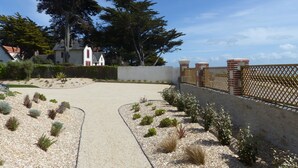Un chemin mène à une maison à l’extérieur blanc, agrémentée de touches de rouge, et qui est entourée d’un jardin de gravier où poussent différentes espèces d’arbustes, le tout délimité par une clôture en bois.