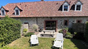 Une maison en pierre avec un toit recouvert de tuiles rouges, deux chaises de jardin blanches et une table de patio.