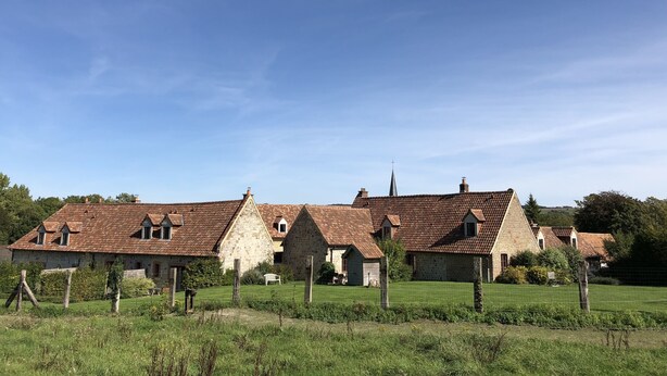 Une rangée de maisons traditionnelles en pierre, coiffées de toits en tuiles rouges, entourées d’un champ verdoyant et surplombées par un ciel bleu clair.