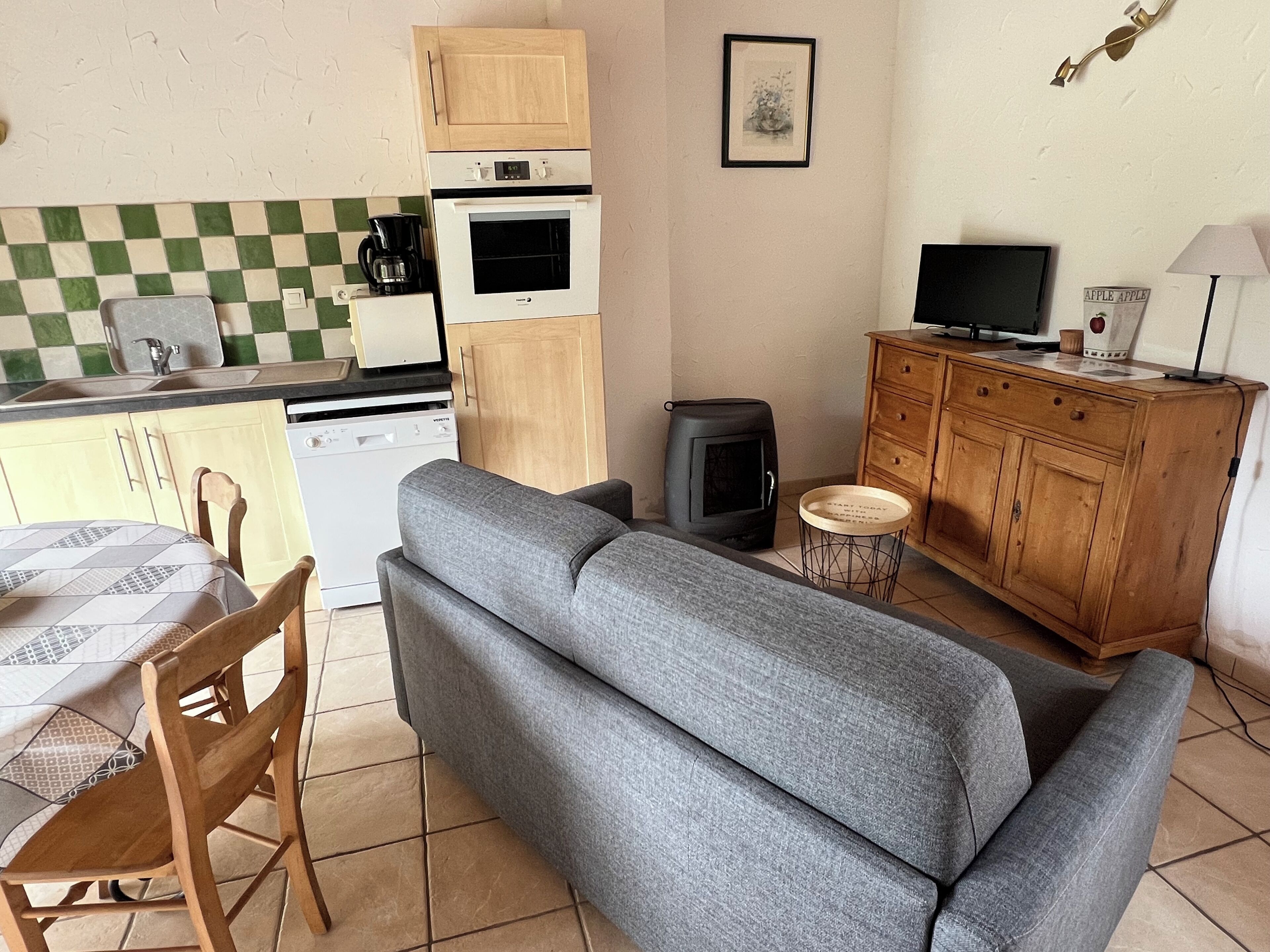 A compact living space with a sofa, a dining table with chairs, a kitchenette with a stove and oven, a television, and a small cabinet.