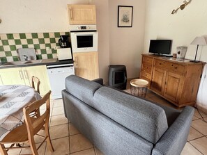 A compact living space with a sofa, a dining table with chairs, a kitchenette with a stove and oven, a television, and a small cabinet.