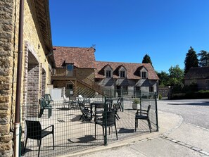 A stone building with a red-tiled roof, outdoor seating, and a fenced patio area.