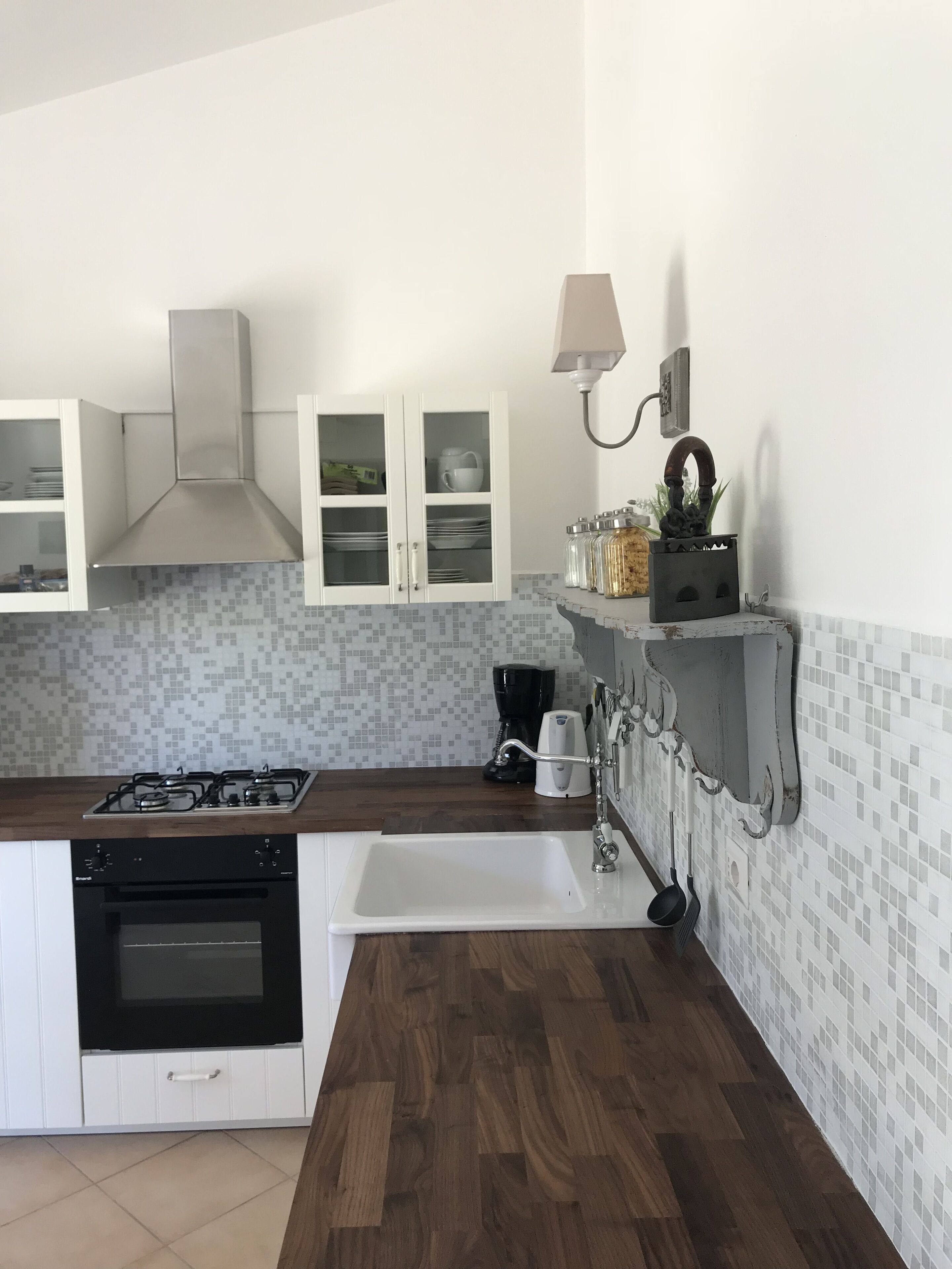 Cocina con estufa a gas, horno, armarios blancos, encimera de madera, salpicadero azulejado, un lavabo y una repisa con frascos y una lámpara.