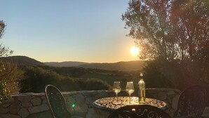 A patio with a table and chairs, two wine glasses, and a bottle, set against a scenic backdrop of hills and a setting sun.