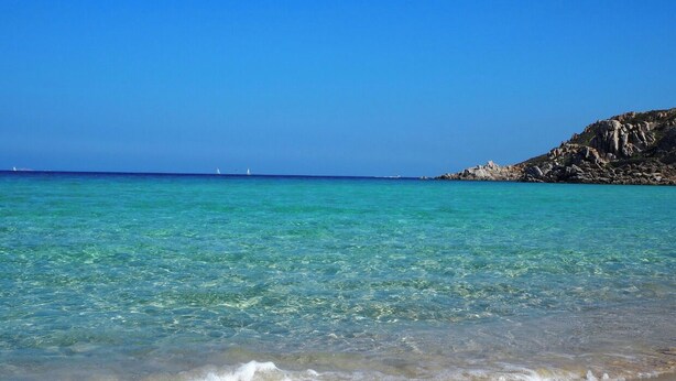 A beach with clear turquoise water, gentle waves, and a rocky outcrop in the background.