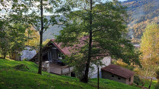 Una cabaña rústica con terraza de madera y techo rojo, rodeada de exuberante vegetación y un paisaje montañoso.