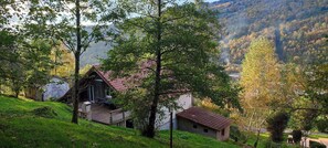 A rustic cabin with a wooden deck and a red roof, surrounded by lush greenery and a mountainous backdrop. - GITE 2 à 20 pers A PARTIR DE 572€/we - WIFI FIBRE GRATUIT - Commerces sur place (Cornimont)