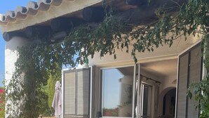 En udendørs terrasse med et rundt bord og fletstole, omgivet af plantede bede og en pergola.