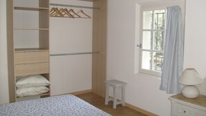 Ein Schlafzimmer mit einem Bett, einem Holzschrank, einem kleinen Hocker, einem Fenster mit Vorhängen und einem Nachttisch mit Lampe.