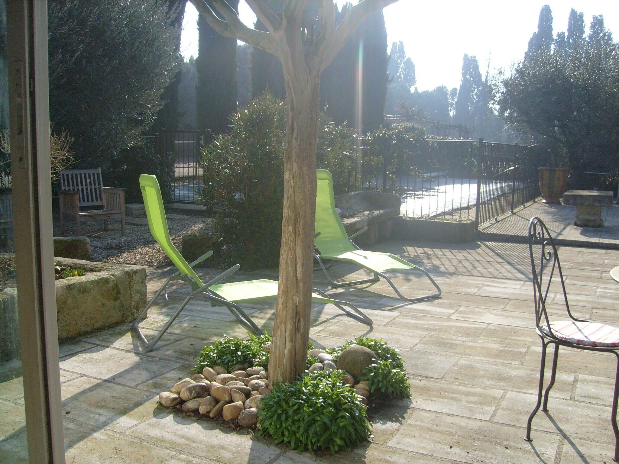 Un patio al aire libre con suelo de piedra, dos sillones verdes, un banco de madera y una silla de metal.