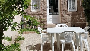 Ein Steinhaus mit einem dreieckigen Dach, einem zentralen Rundbogenportal und einer Schotterterrasse mit weißen Kunststoffstühlen.