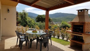 Eine Terrasse im Freien mit einem runden Tisch und Stühlen, einem Backofen aus Ziegelsteinen und Blick auf Berge.