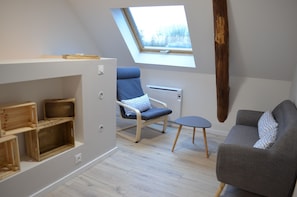 Une pièce avec un plafond en pente, une lucarne, une étagère en bois, une chaise bleue, un canapé gris et une petite table.
