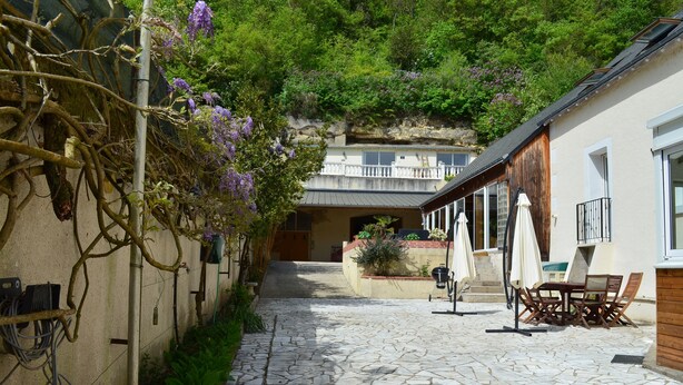 A stone-paved courtyard with a wooden fence, outdoor seating, and a view of a building with a balcony. - Gîte de la Merluchette  (Nazelles-Négron)