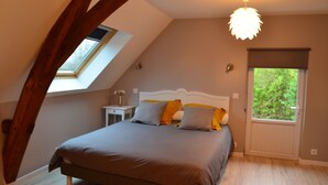 A bedroom with a sloped ceiling, a bed with grey and yellow bedding, a bedside table with a lamp, and a window with a view of greenery. - Gîte de la Merluchette  (Nazelles-Négron)