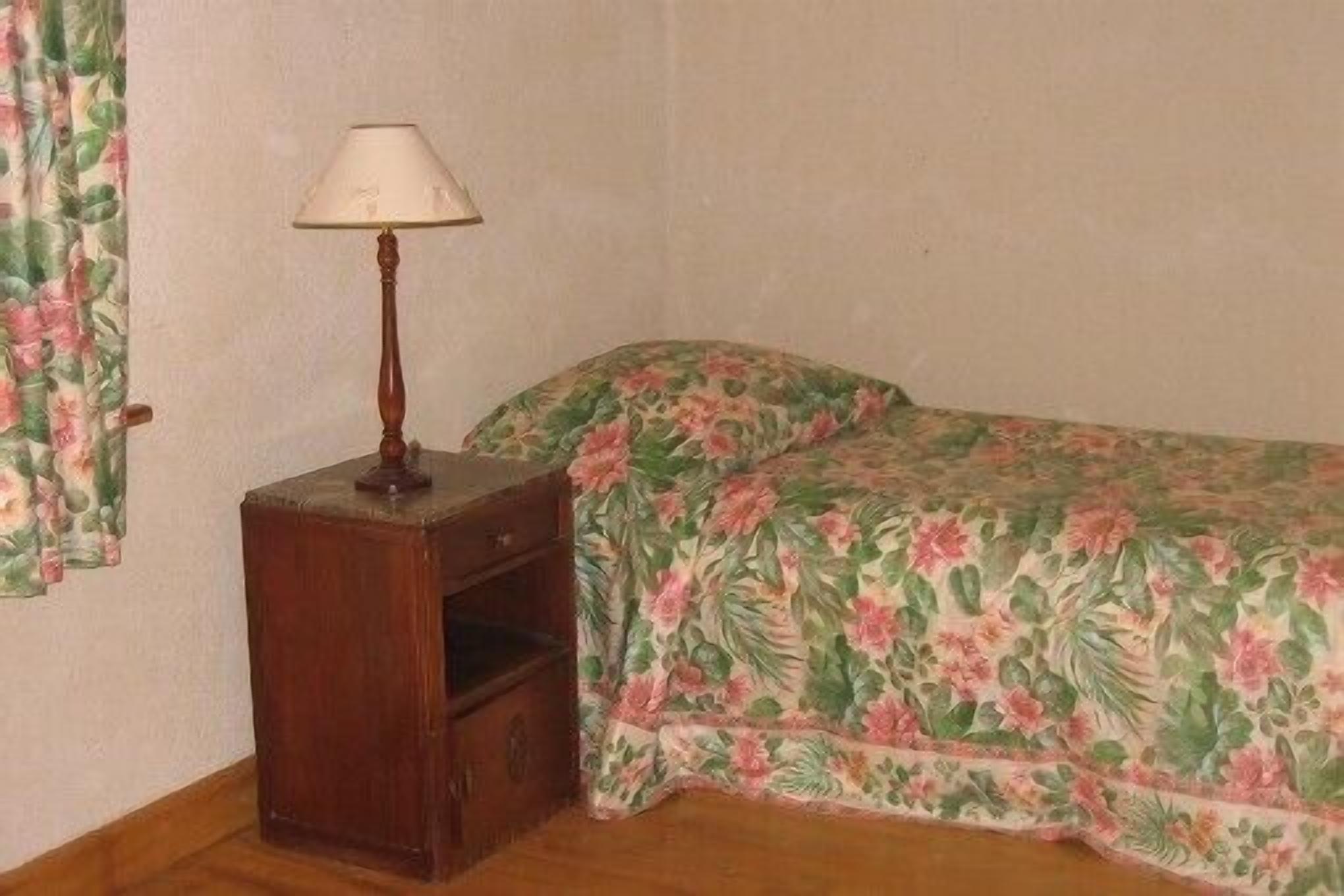 Ein kleines Zimmer mit Nachttisch aus Holz, einer Lampe, einem Bett mit Blumenmuster auf der Bettwäsche und einem Fenster mit Vorhängen.
