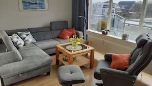 A modern living room with a grey sofa, a matching armchair, and a small table with a vase of flowers. A window provides a view of buildings and a street outside. -  Bright and friendly apartment in the center of Grömit (Grömitz)