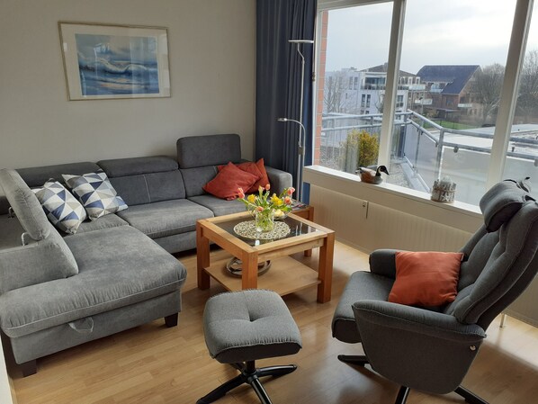 A modern living room with a grey sofa, a matching armchair, and a small table with a vase of flowers. A window provides a view of buildings and a street outside. - Bright and friendly apartment in the center of Grömit (Grömitz)