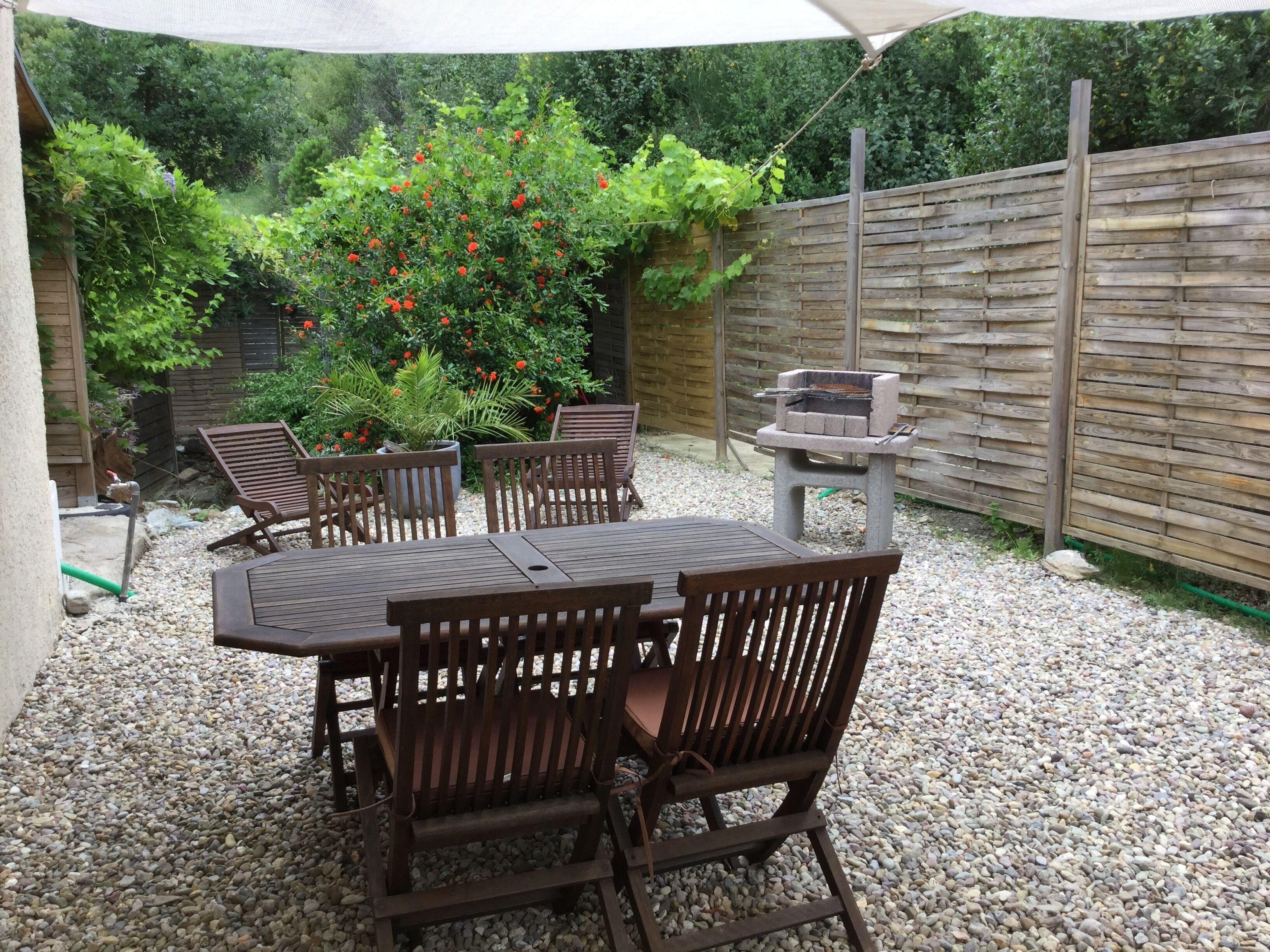 Eine Terrasse mit Holztisch und -stühlen, einem Grill und einem Garten mit verschiedenen Pflanzen und einem Holzzaun.