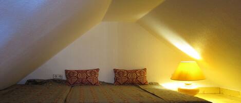 A small, cozy bedroom with a sloped ceiling, a single bed with floral bedding, two decorative pillows, and a bedside lamp.