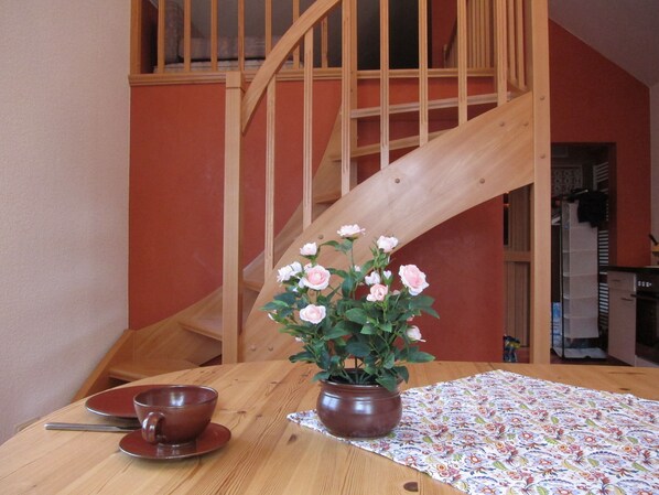 Una escalera de madera con barandilla de madera, una mesa de comedor con mantel floral, una maceta de cerámica marrón con flores blancas, y una taza y platillo marrones.