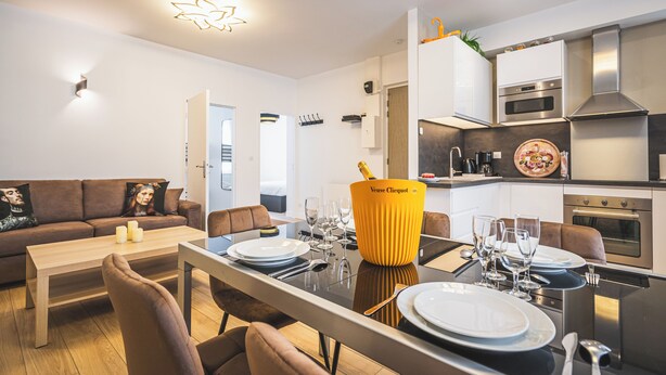 Une cuisine moderne équipée d’appareils électroménagers en acier inoxydable, un coin repas avec une table dressée pour quatre personnes et un salon avec un canapé et des coussins.